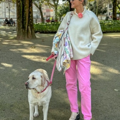 Mulher com camisola branca, calças rosas e botas brancas a passear cão labrador branco num parque.