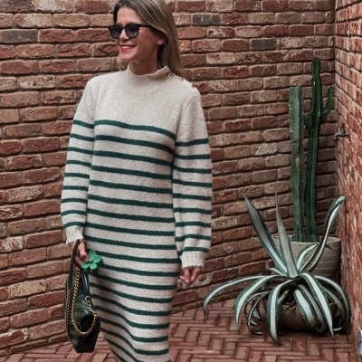 Mulher com vestido de malha riscada verde e branco, óculos escuros, mala preta, parede de tijolo e plantas