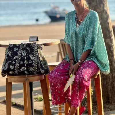 Mulher com roupa colorida numa esplanada à beira-mar com mala preta e barco ao fundo.