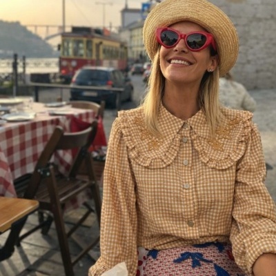 Mulher com chapéu de palha e óculos de sol vermelhos sentada numa mesa de restaurante com toalha xadrez