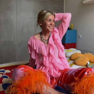 Pessoa num quarto com roupa rosa e calças vermelhas com plumas, colares, óculos azuis no chão e cama com edredão vermelho