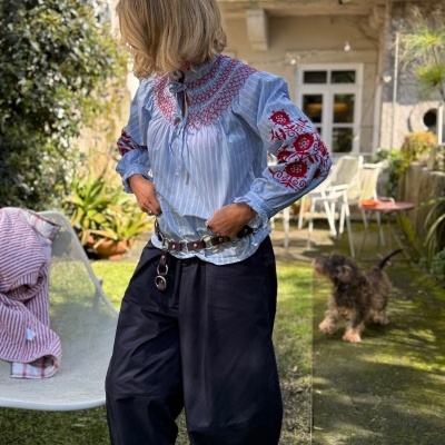 Mulher com blusa bordada azul e calças largas pretas num jardim