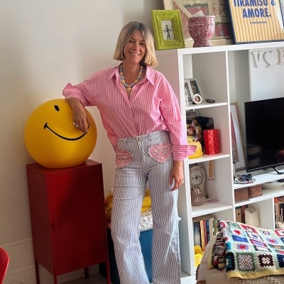 Mulher com camisa rosa às riscas e calças cinza às riscas, colar colorido, junto a baú vermelho com bola amarela sorridente em sala luminosa