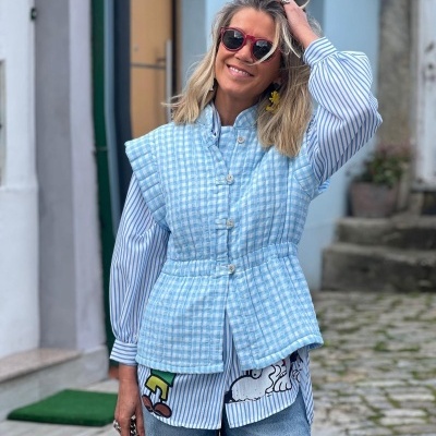 Mulher vestindo colete azul claro xadrez, camisa azul e branca riscada com estampa Snoopy, calças de ganga e óculos de sol vermelhos numa rua com calçada de pedra.