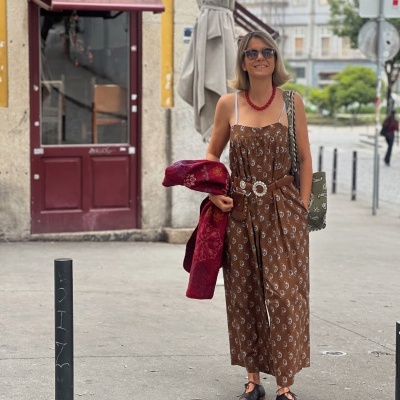 Mulher com vestido castanho floral, cinto branco, colar vermelho e óculos escuros caminhando na rua.