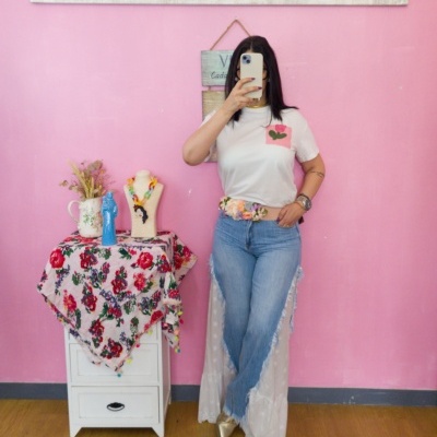 Mulher com t-shirt branca, jeans azuis e cinto de flores tirando selfie em espelho, junto a gaveteiro com toalha floral e decoração