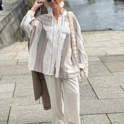 Mulher com blusa às riscas, calças largas creme, sapatilhas brancas e bolsa trançada junto a rio