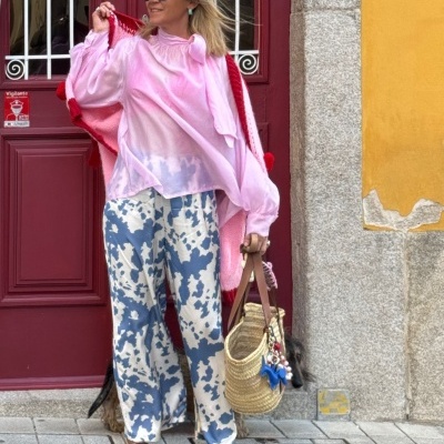 Mulher com blusa rosa, pantalones estampados azuis e brancos, sapatos brancos e marrons, bolsa de palha em frente a porta vermelha