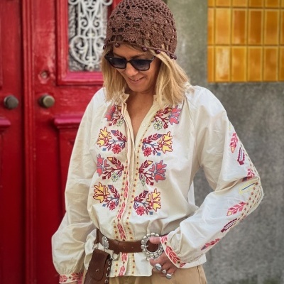 Blusa branca bordada colorida com cinto castanho, chapéu castanho de crochê e óculos escuros, mulher em ambiente urbano.