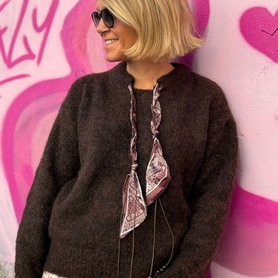 Mulher de cabelo loiro com camisola castanha felpuda junto a parede rosa com grafite.