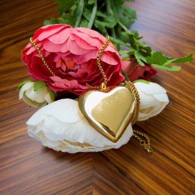 Colar dourado em forma de coração sobre flores branca e rosa em madeira clara