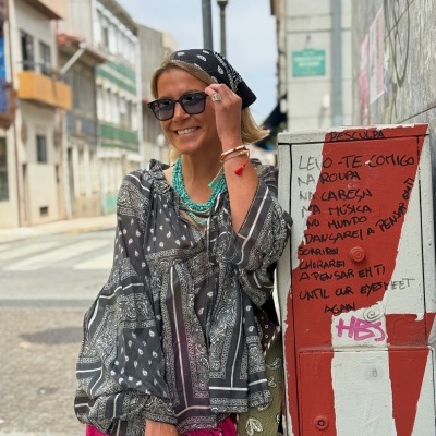 Mulher com blusa e lenço pretos com padrão paisley branco, calças rosa com padrão paisley, colares turquesa e pulseiras, junto a poste com graffiti e texto em rua urbana