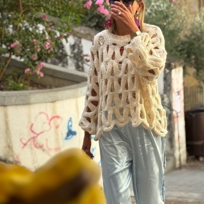 Pessoa com camisola de malha creme de laços grandes e calças cinza numa rua com flores cor-de-rosa.