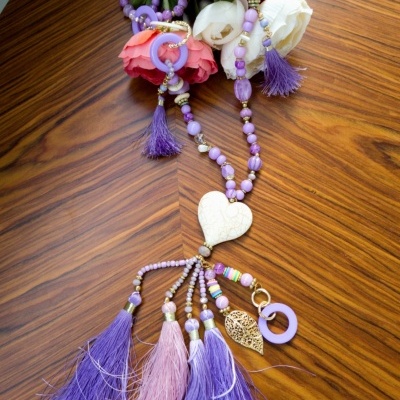 Colar com contas lilases, pingentes em forma de coração, borlas coloridas sobre mesa de madeira e flores artificiais