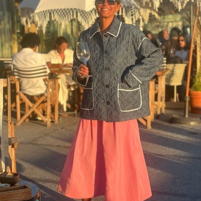 Mulher com casaco acolchoado cinzento, vestido rosa e sapatos amarelos segura copo em esplanada com guarda-sóis brancos