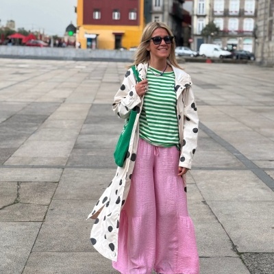 Mulher com casaco branco de bolas pretas, blusa verde às riscas, calças rosa largas e sapatilhas num espaço urbano