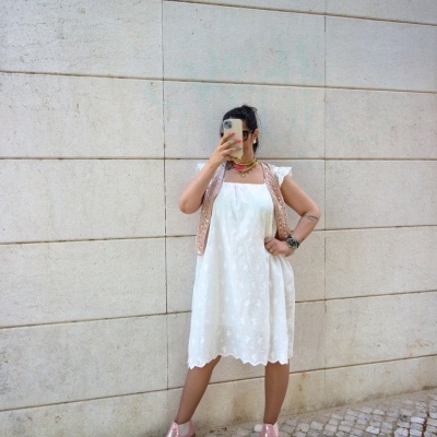 Mulher de vestido branco, colete bege, botas cor de rosa, posando contra parede branca