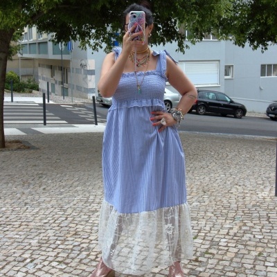 Mulher com vestido azul às riscas e renda branca abaixo, sandálias rosa dourado, tirando selfie num passeio de pedra com árvores e carros ao fundo