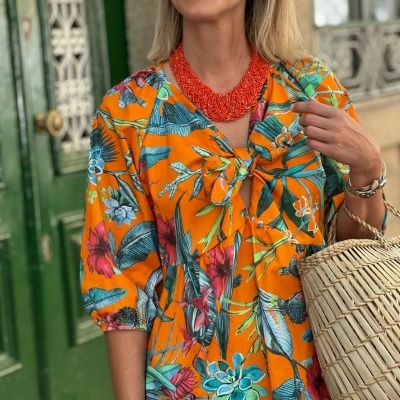 Mulher com vestido floral laranja, colar vermelho, bolsa de palha, óculos de sol e porta verde com ferragens em fundo