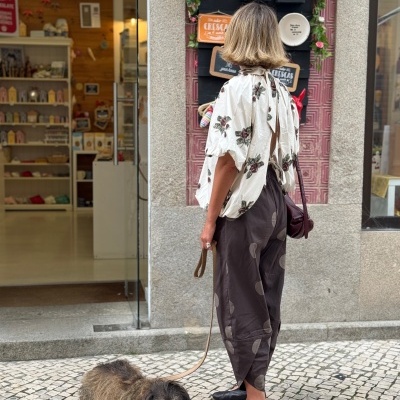 Mulher de costas com blusa floral e calças castanhas com cão de trela no chão em frente a loja decorada.