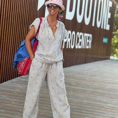 Mulher com roupa clara de riscas e flores, chapéu rosa, óculos, mochila azul e saco vermelho em passadeira de madeira junto a painel DUOTONE PRO CENTER.