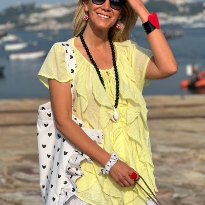 Mulher com blusa amarela e saia branca junto ao mar, acessórios coloridos e bolsa branca com corações pretos.