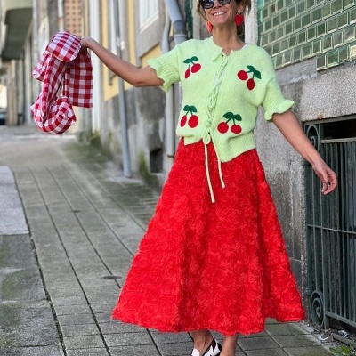 Mulher com casaco verde claro com cerejas, saia vermelha texturizada e sapatos preto e branco numa rua de calçada
