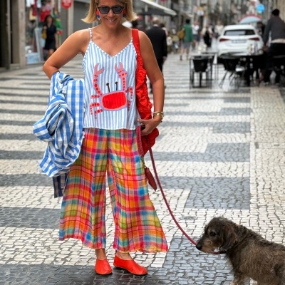 Mulher com top de alças com caranguejo, calças largas xadrez, sapatilhas vermelhas e lenço amarelo na cabeça numa rua calçada