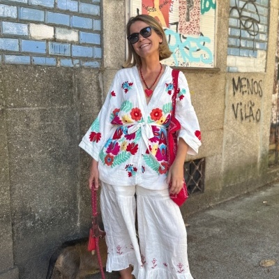 Mulher sorridente de camisa e calças brancas bordadas junto a parede com azulejos azuis e grafiti