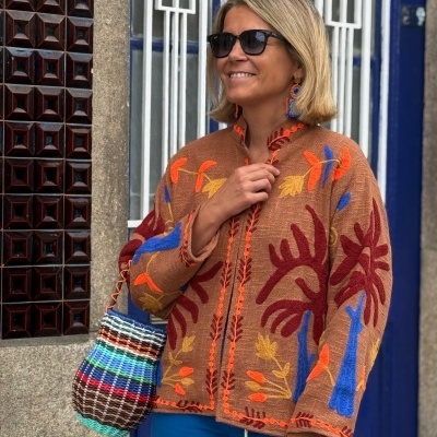 Mulher com casaco castanho bordado, calças azuis e saco de palha colorido a sorrir junto a porta azul