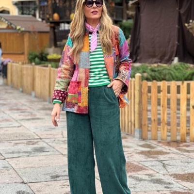 Mulher com casaco colorido de patchwork, t-shirt às riscas, calças verdes largas e lenço rosa, em zona exterior urbana.
