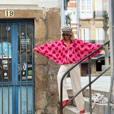 Mulher com casaco rosa com corações vermelhos, calças brancas com corações, sapatos vermelhos e chapéu listrado numa escadaria ao lado de porta azul.