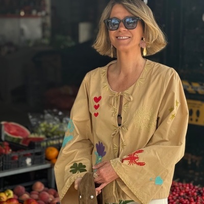 Blusa bege bordada com motivos coloridos num mercado com frutas e caixas ao fundo.