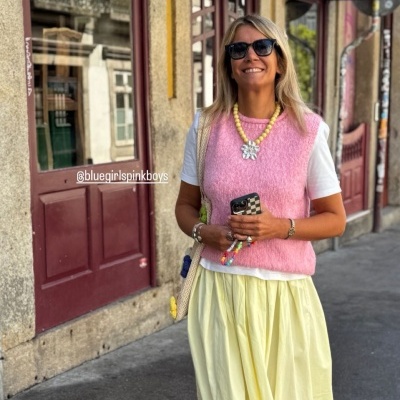 Mulher vestindo colete rosa, saia amarela e t-shirt branca na rua com parede amarela ao fundo