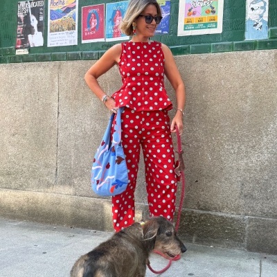 Mulher com conjunto vermelho de bolas azuis e cão com trela vermelha numa rua urbana com cartazes coloridos no muro verde.