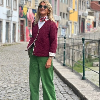 Mulher com casaco borgonha, calças verdes e sapatos beges em rua de calçada