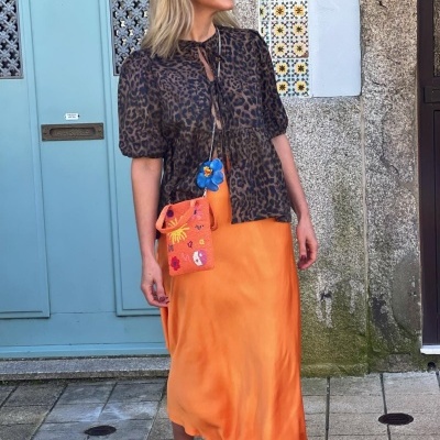 Mulher com blusa animal print, saia laranja, sapatos azuis e bolsa bordada em frente a porta azul com cerâmica