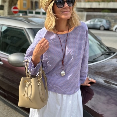 Mulher com chapéu de palha, óculos escuros pretos, blusa xadrez azul e rosa, saia branca, bolsa bege em pele, junto a carro preto na rua.