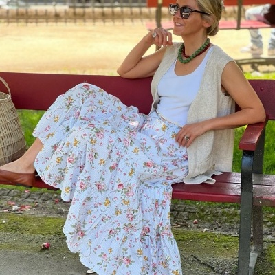 Mulher sentada num banco vermelho usando saia floral, blusa branca e colete bege com óculos de sol.