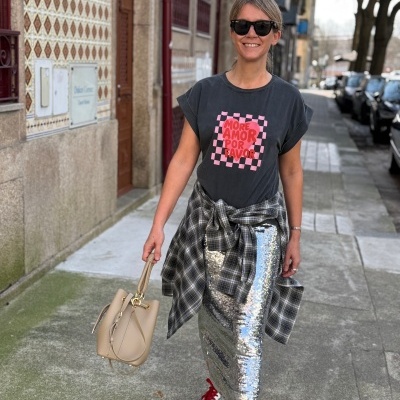 Mulher com t-shirt preta com texto vermelho, saia de lantejoulas prateada, camisa xadrez amarrada na cintura e mala bege