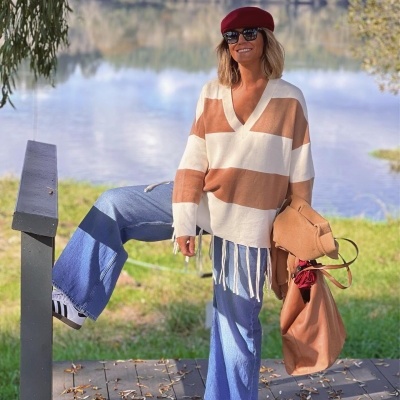 Mulher com casaco de malha riscas beges e brancas, calças jeans largas, ténis brancos, óculos escuros e boina vermelha junto a lago com árvores