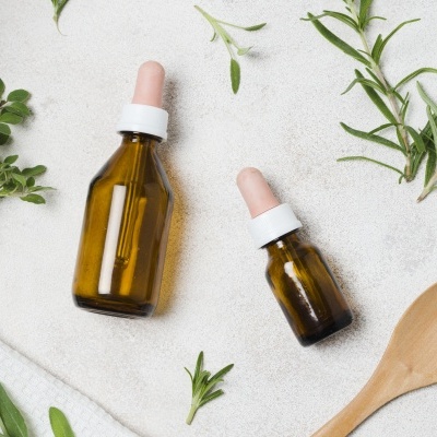 Frascos de vidro âmbar com conta-gotas rosa com folhas verdes e colher de pau em fundo claro