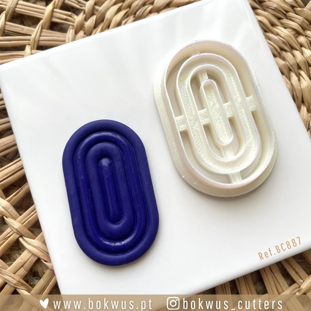Cortador Geométrico (5cm) - Versão Sólida e Vazada Dois cortadores ovais, azul escuro e branco, com padrão geométrico em relevo, sobre placa branca em fundo de vime.