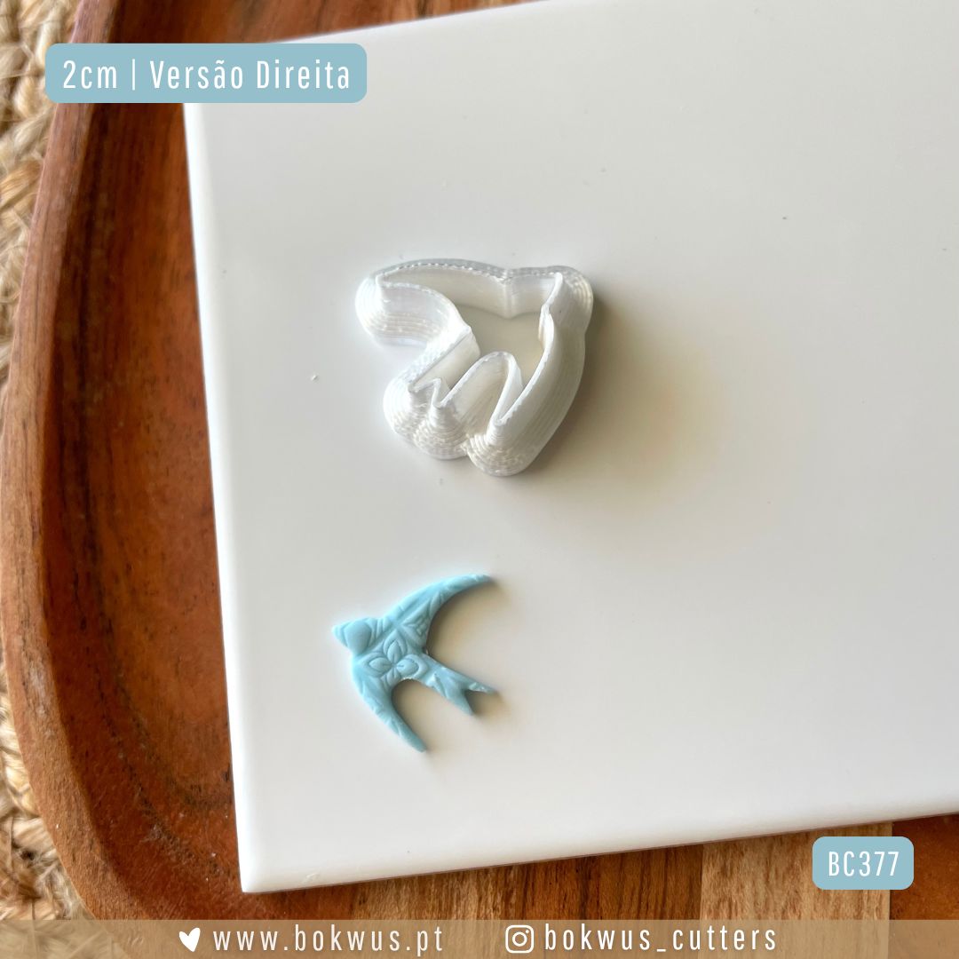 Cortador Andorinha | Disponível em 5 tamanhos Cortante de metal branco em forma de pássaro com texto de tamanho 2cm num prato branco sobre base de madeira.