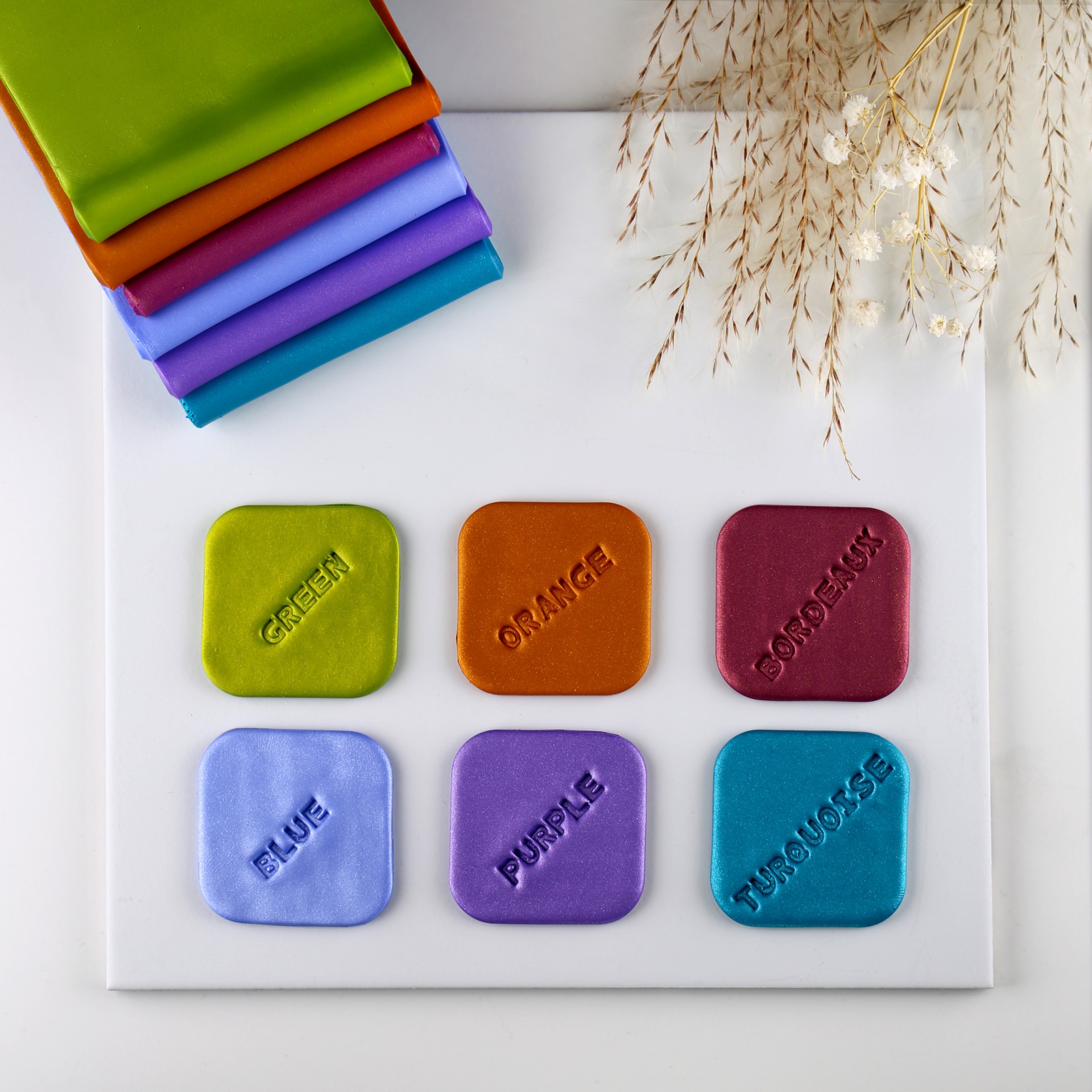 Amostras quadradas de tecido colorido com texto em relevo sobre placa branca, com tecidos dobrados coloridos e ramos secos ao lado.