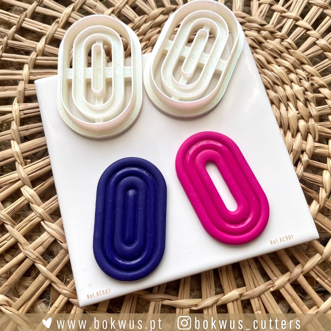 Cortador Geométrico (5cm) - Versão Sólida e Vazada Quatro cortadores de plástico em forma oval com relevo, dois brancos, um azul e um rosa, sobre base branca em tecido de rattan.