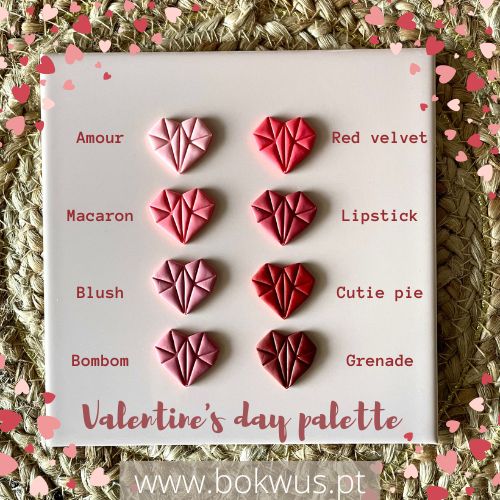 Paleta de maquilhagem com sombras em forma de coração em cores rosa e vermelho, nomes das cores escritos ao lado, sobre tecido trançado bege