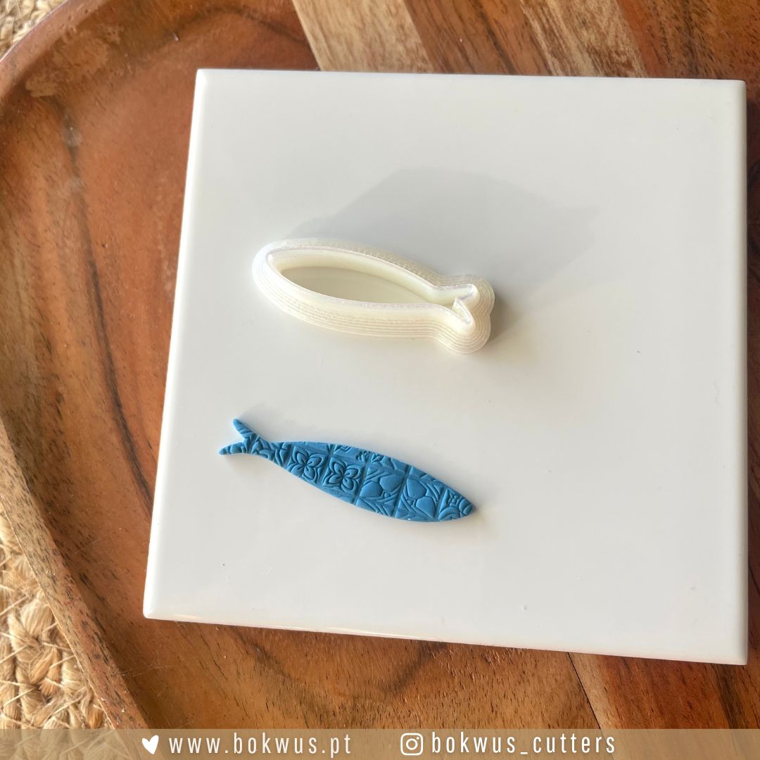 Cortador Sardinha | Disponível em 4 tamanhos Dois cortadores para confeitaria, um branco em forma de laço e um azul em forma de peixe com texto 'BKWUS', sobre placa branca em madeira.