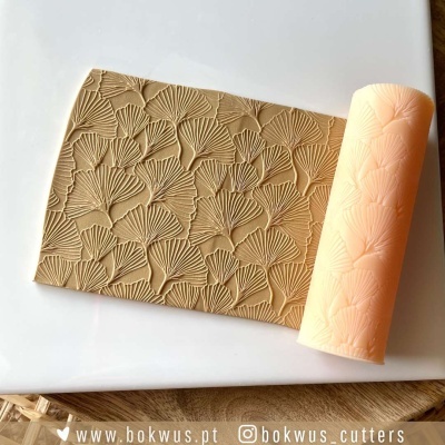 Rolo de textura branco com padrão de folhas e peça de massa bege com o mesmo padrão sobre superfície branca