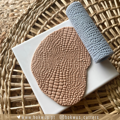 Rolo texturizador para argila com padrão escamado azul claro e argila bege moldada com o mesmo padrão sobre fundo de fibra natural.